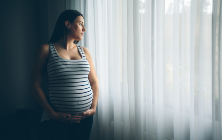 An expectant mother stares out the window as she holds her pregnant belly. 