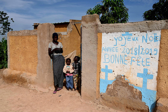 A woman and children stand at a fence with mural reading 'Merry Christmas 2018/2019 - happy celebrations' in the village of Yagma, north of Ouagadougou, on September 17, 2019. Burkina Faso, a poor country in West Africa, has been caught in a spiral of violence attributed to jihadist armed groups that has led to the closure of schools in the north and east of the country. Jihadist attacks have internally displaced more than 300,000 people including many children who have dropped out of school. 