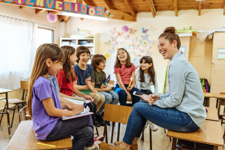 A teacher sits on top of a desk in front of several students.