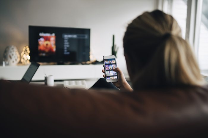 Rear view of teenage girl using smart phone app while watching TV in living room at home 