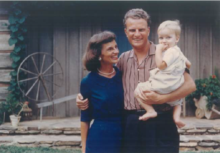 Ruth and Billy Graham smile in front of their mountain home in Montreat, holding their son Franklin.