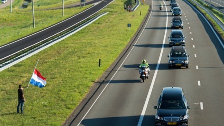 A man holds a Netherlands flag flying at half-mast as a row of hearses, escorted by military police, passes by.