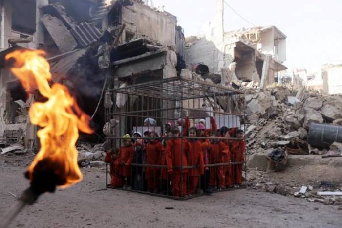 An activist (not pictured) holds a burning torch near children carrying banners inside a cage during a protest against forces loyal to Syria's President Bashar al-Assad in Douma, eastern Al-Ghouta, near Damascus February 15, 2015.