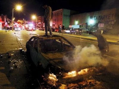 A rioter stands atop a burning car as another man pours fuel onto the fire while Baltimore firefighters behind them fight fires in mutliple burning buildings set ablaze by rioters during clashes in Baltimore, Maryland April 27, 2015.